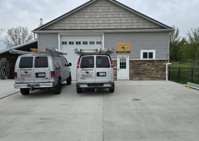 Two white vans with company logos are parked in front of a large gray building with a stone accent and a garage door. The building has a sign above the door and is surrounded by a concrete lot.