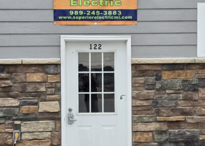 A white door labeled 122 is flanked by stone and gray siding. Above it is a sign for "Superior Electric" with a phone number and website URL.