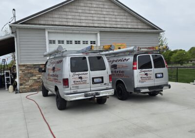 Two white Superior Electric vans parked in front of a gray building with stone accents. One van's rear door is open, and equipment is visible. A red extension cord is stretched across the driveway.